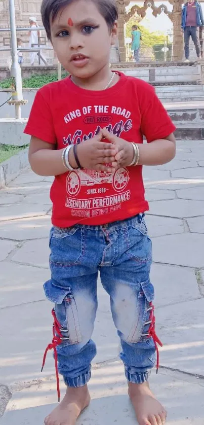 Young child in a red shirt and blue jeans poses outdoors.