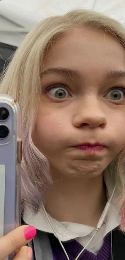 Young girl takes a selfie with playful expression holding a smartphone.