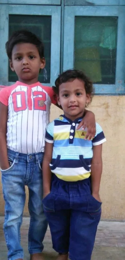 Two young children standing by a blue window, exuding charm and style.