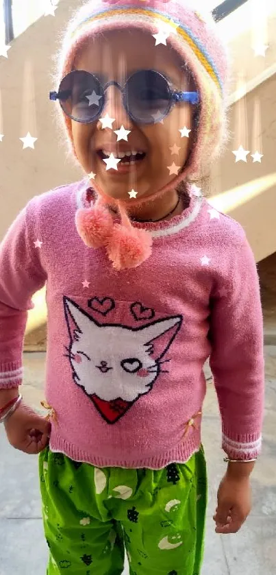 Smiling child in a pink cat sweater with green pants and sunglasses.