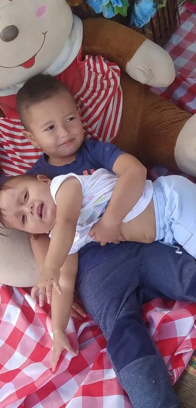 Two toddlers enjoy a picnic with a large teddy bear on a checkered blanket.