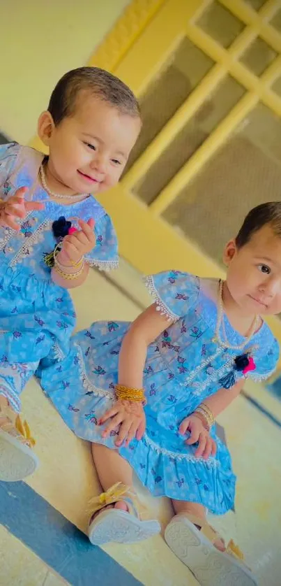 Cute twin toddlers in blue dresses sitting indoors.