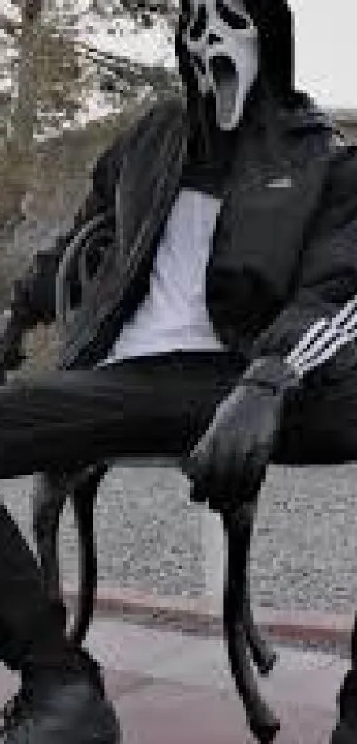 Masked figure in black jacket sitting outdoors on a chair.