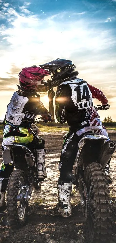 Two motocross riders sharing a moment at sunset on a dirt track.