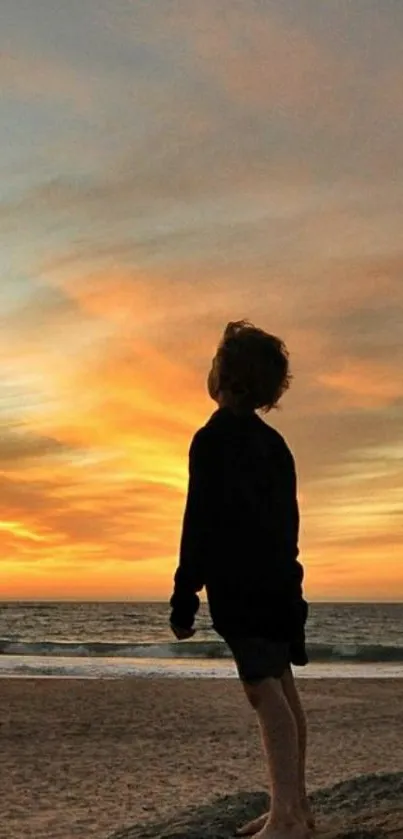 Child standing on the beach during a vibrant orange sunset, gazing at the ocean.