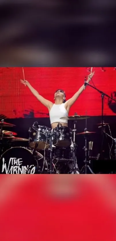 Drummer performing energetically on stage with a red backdrop.