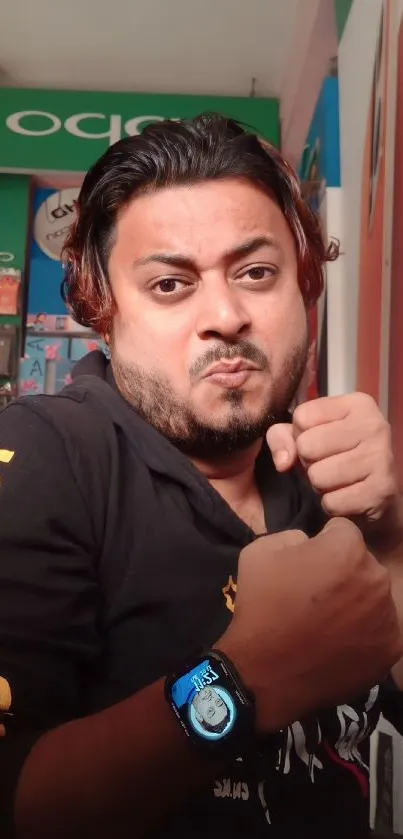 Man posing in a tech store with gadgets and poster backdrop, wearing a watch.