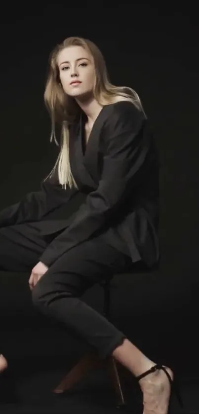 Elegant person in a black suit against a dark background, seated stylishly.