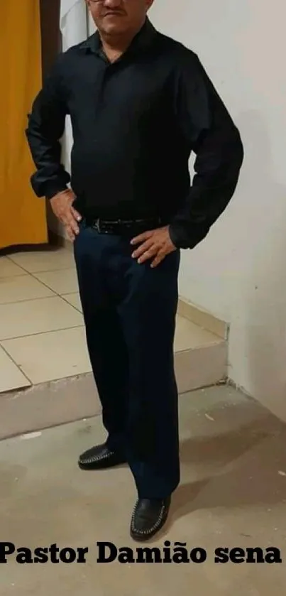 Man in black attire posing against beige wall.