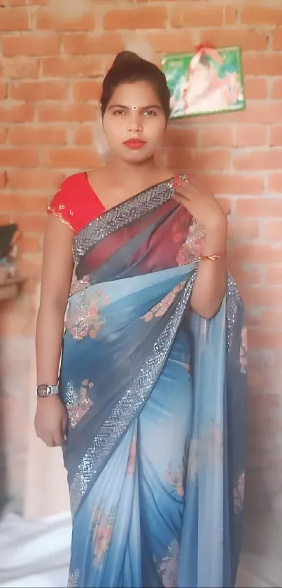 Woman in a blue saree standing against a brick wall.