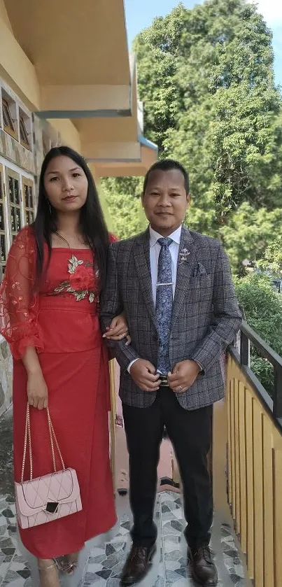 Elegant couple standing on a balcony with lush green background.