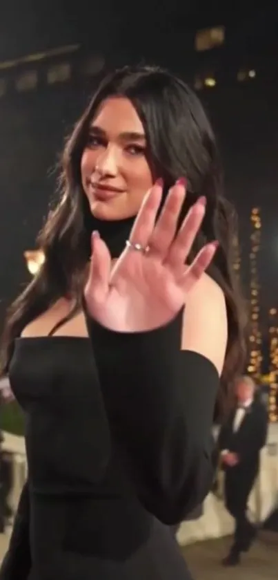 Elegant woman in black dress, waving at a nighttime event.