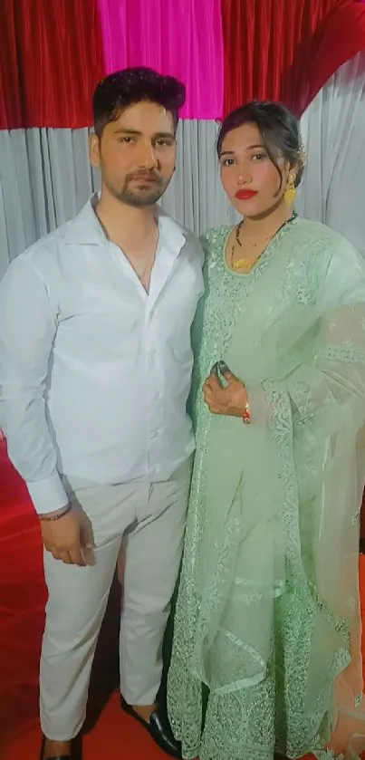 Elegant couple in traditional attire at a festive event with vibrant red backdrop.