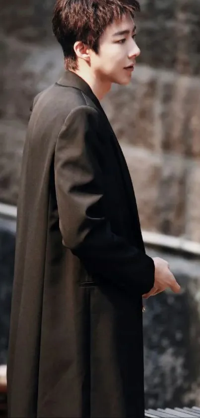 A man in a long black coat stands by a stone wall, exuding elegance.