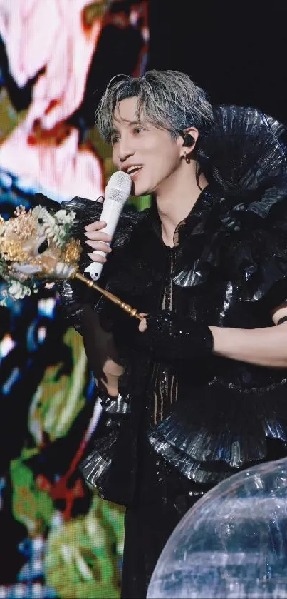 Performer in black outfit holding microphone on stage with a floral background.