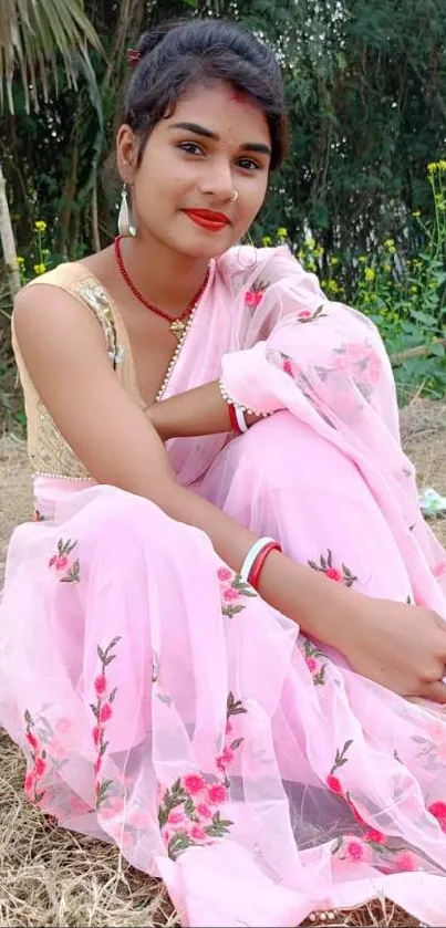 Woman in pink saree sitting outdoors with floral design.