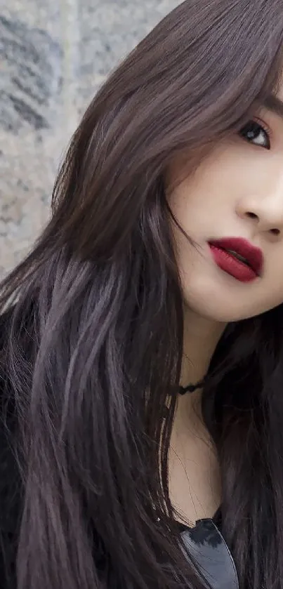 Portrait of a model with dark hair and red lips against a stone background.