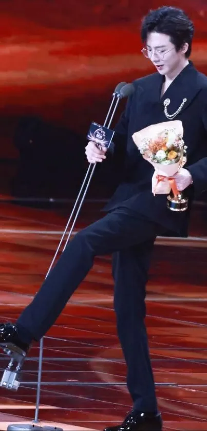 Person in elegant black suit holding flowers on stage.