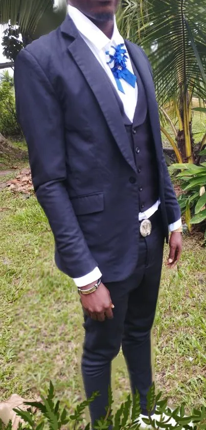 Man in dark suit and blue tie in lush greenery.