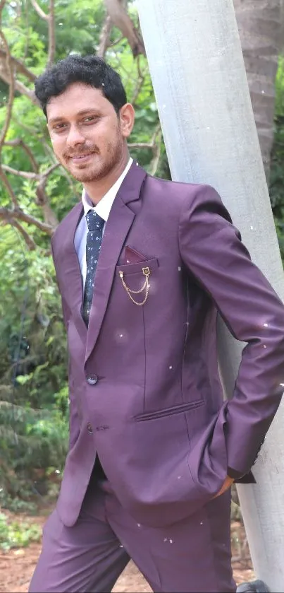 Man in a burgundy suit leaning against a pole with lush green background.