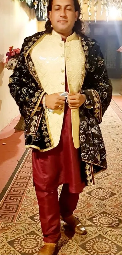 Man in elegant traditional attire with ornate jacket and burgundy robe on patterned rug.