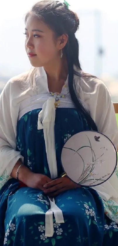 Woman in traditional dress sitting with a decorative fan, in an outdoor setting.