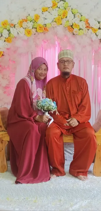 Couple in traditional attire seated against a floral backdrop.