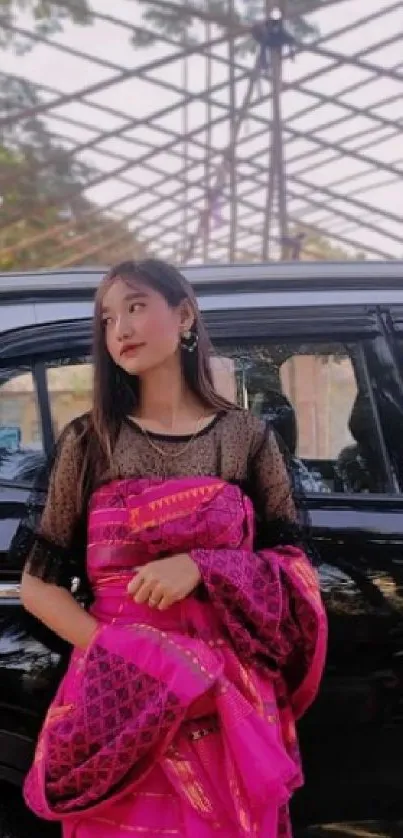 Woman in pink sari stands by a black car in sunlight.