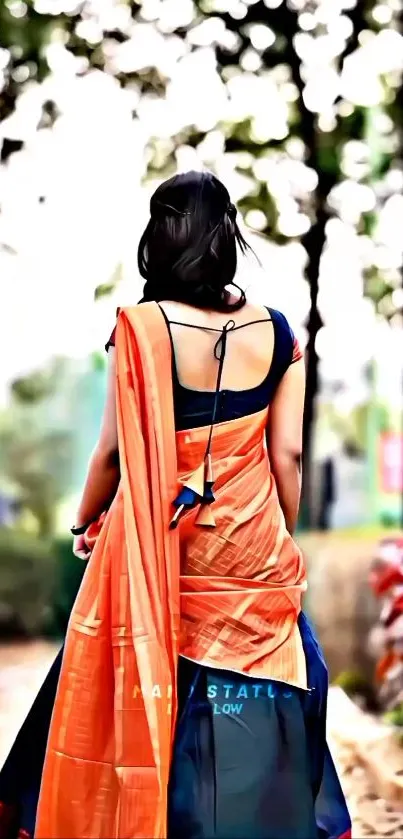 Woman in an orange saree walking in a garden path with a nature backdrop.