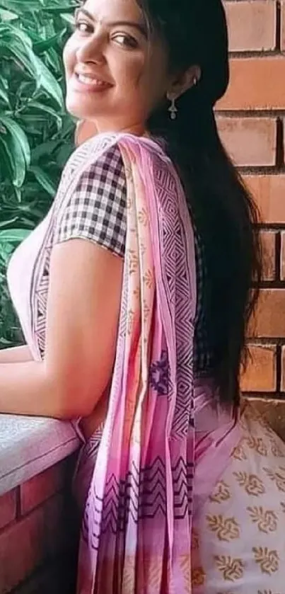 Woman in pink patterned sari smiling by a brick wall with greenery.