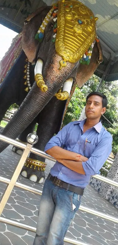 A man in blue shirt standing beside a large decorated elephant statue outdoors.