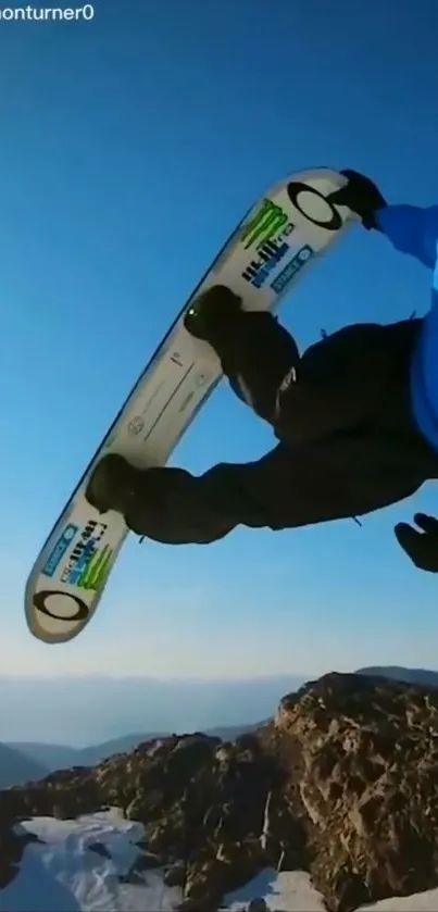 Snowboarder in mid-air against a mountain backdrop under a blue sky.