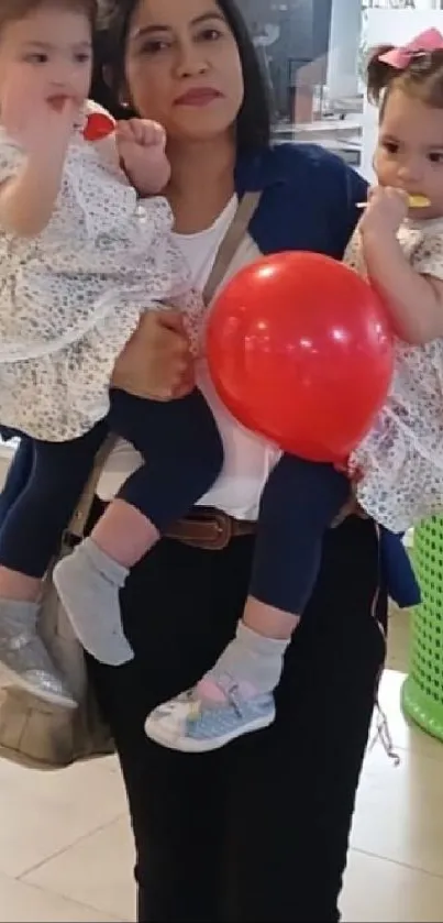 Mother holding twins with a red balloon in a mall setting.