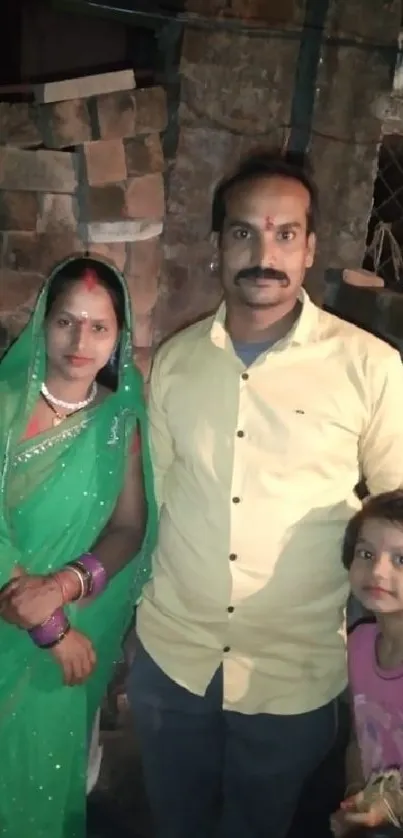 A family in traditional attire poses by a brick wall, with the woman in a green sari.