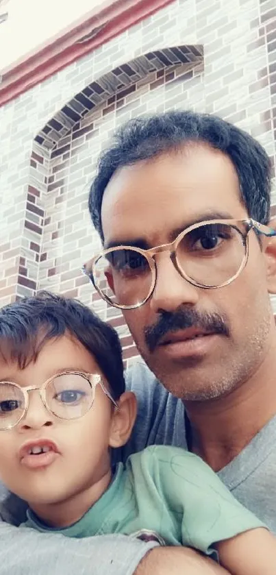 A father holding his child close, both wearing glasses, with a brick wall background.