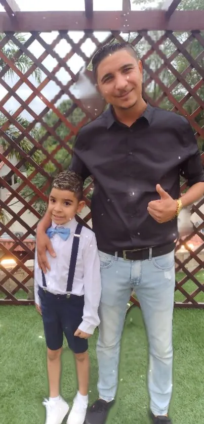 Father and son in stylish outfits near a garden fence.