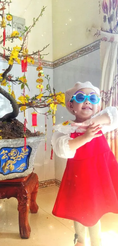 Child in red dress beside festive bonsai tree in sunlit room.