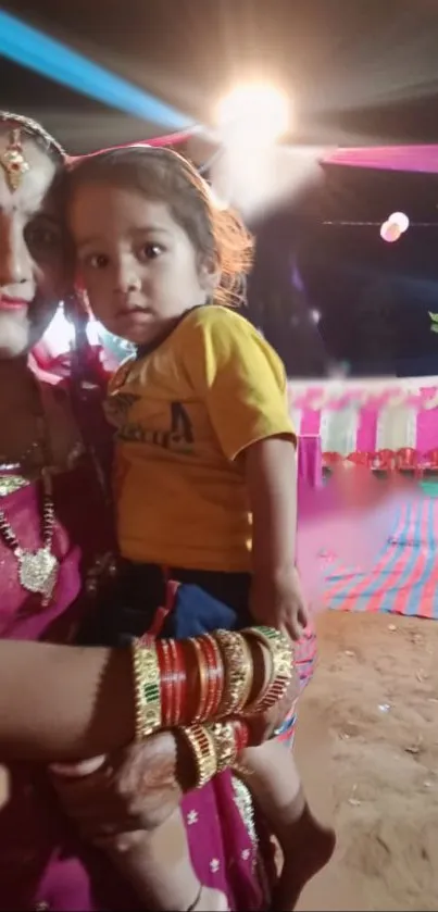 A woman in traditional attire holding a child during a festive evening celebration.