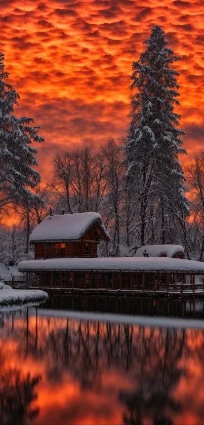 Snow-covered cabin by lake with fiery sunset sky reflecting in water.