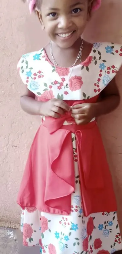 Smiling girl in floral dress with red sash, standing against a pink textured wall.