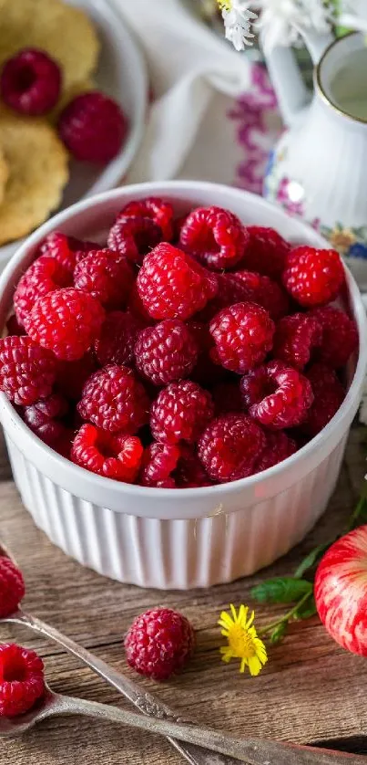 Bowl of red raspberries with flowers and apples on wooden table.