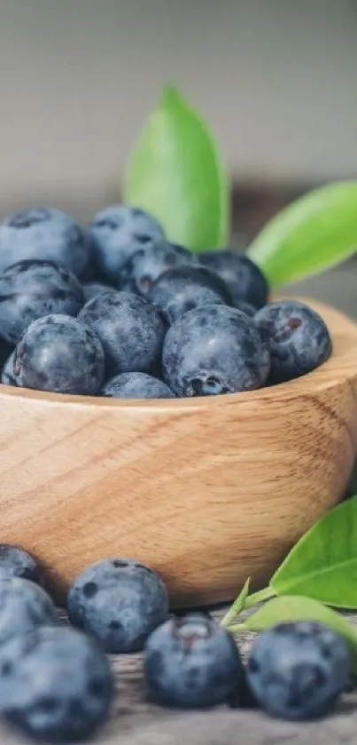 A wooden bowl filled with fresh blueberries and green leaves on rustic wood surface.