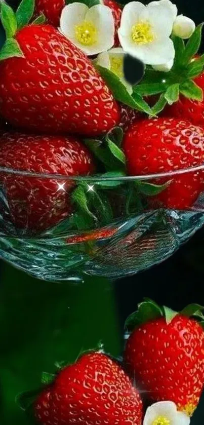 Bright strawberries with flowers in a clear glass bowl on a dark background.