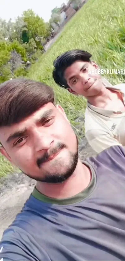 Two friends taking a selfie in a sunny, green field with clear skies.