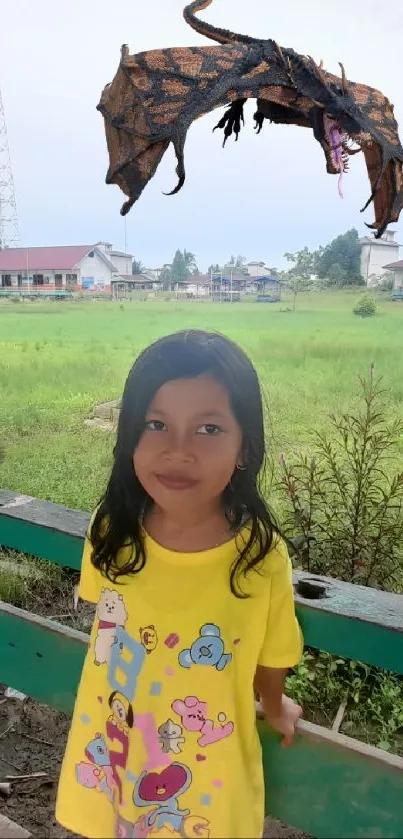 Girl in a yellow dress with a dragon in a countryside setting.