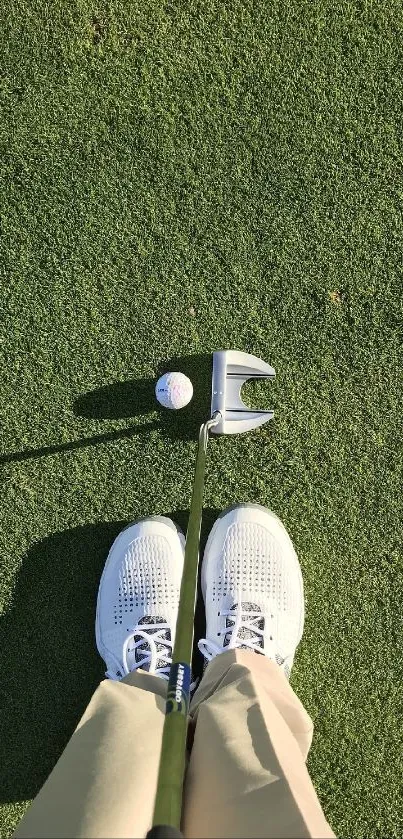 Golf ball and putter on green grass with shoes visible.