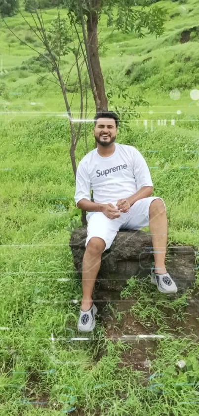 Man sitting on a rock in a lush green field with trees and hills in the background.