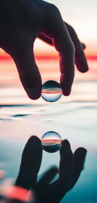 Hand holding a reflective water droplet at sunset on a calm body of water.