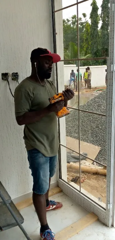 Construction worker with drill by a window amid building site.