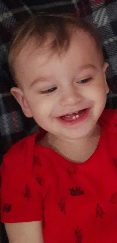 Smiling baby in red shirt with plaid backdrop.